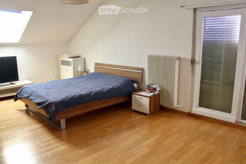 Bright bedroom with wooden flooring, a bed with a blue duvet, a nightstand with a lamp, a radiator, a TV on a stand, and a window with blinds.