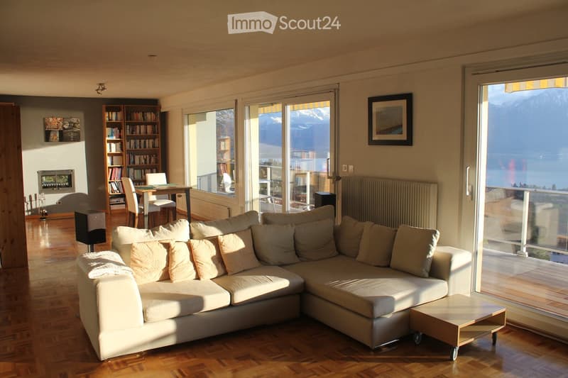 Bright living room with large windows offering a scenic mountain view, featuring a white sectional sofa, a wooden coffee table, a bookshelf, a dining table with chairs, and a fireplace.