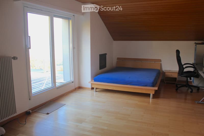 Bright bedroom with wooden flooring, large window, bed with blue bedding, desk with chair, and nightstand.