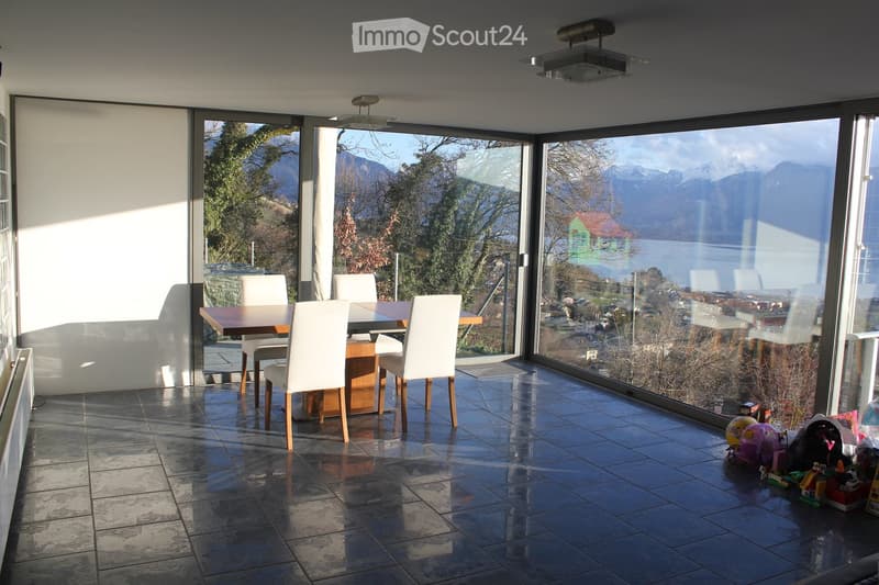 Bright dining area with large windows offering scenic mountain and lake views, featuring a wooden dining table and white chairs.