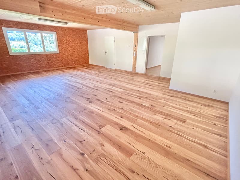 Empty room with wooden flooring, white walls, and exposed wooden beams on the ceiling. Features a large window with multiple panes and several doors leading to other spaces.