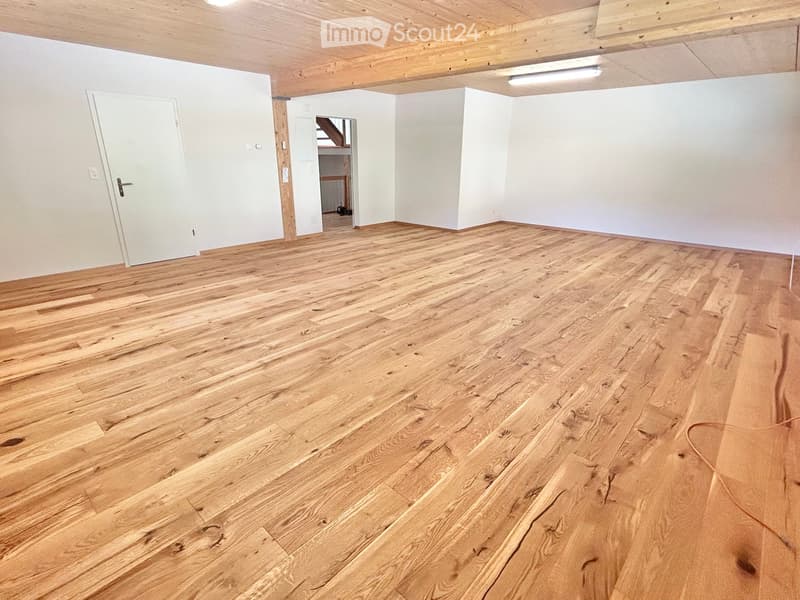 Empty room with wooden flooring, white walls, and exposed wooden beams on the ceiling. Features a white door and a visible staircase leading to another level.