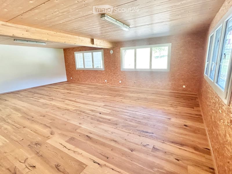 Empty room with wooden flooring, exposed wooden ceiling beams, and multiple windows allowing natural light.
