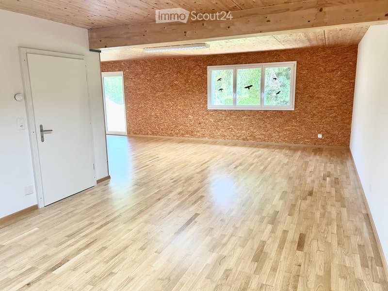 Empty room with wooden flooring, white walls, exposed wooden ceiling beams, and large windows with decorative cutouts.