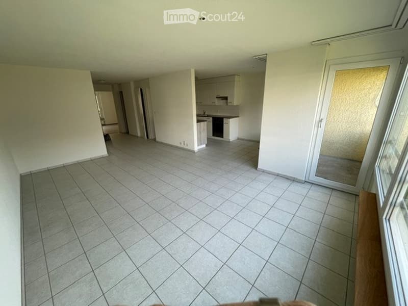 Empty room with tiled floor and white walls, featuring a small kitchen area with cabinets and a sink in the background.