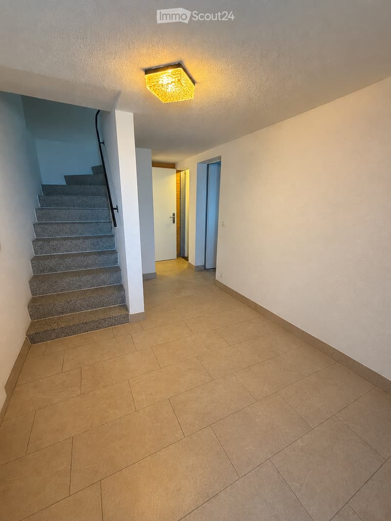 Empty hallway with tiled floor, beige walls, staircase leading upwards, and a bright ceiling light fixture.
