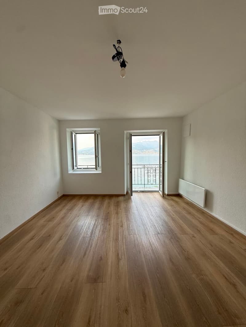 Empty room with wooden flooring, white walls, a window, and an open door leading to a balcony with a view of the sea.