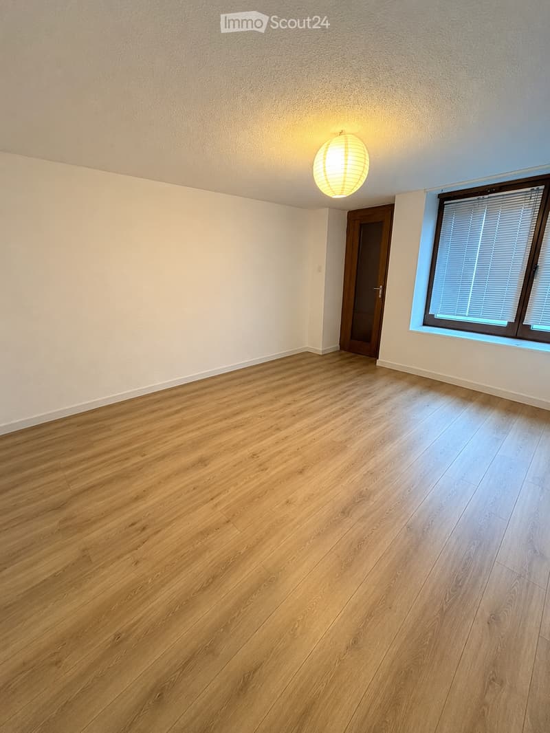 Empty room with wooden flooring, white walls, a single window with blinds, and a door leading to another space.
