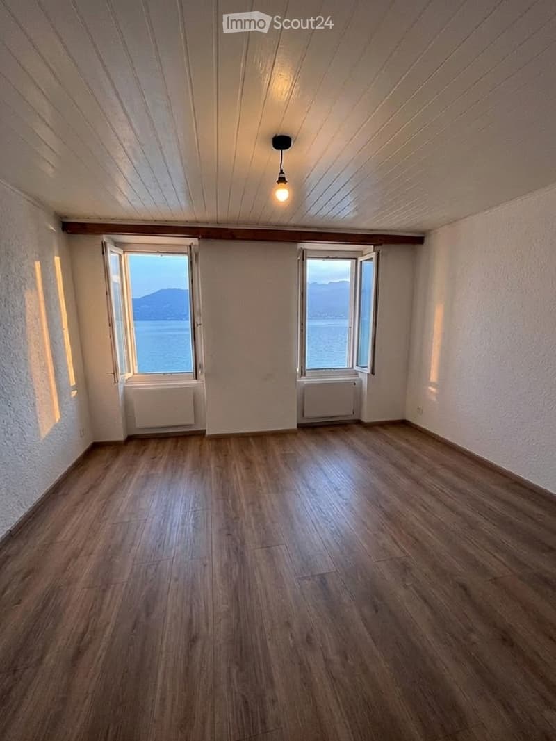 Empty room with wooden flooring, white walls, and large windows offering a scenic view of the sea and mountains.