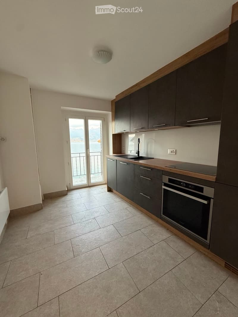 Modern kitchen with dark grey cabinets, a sink, faucet, and built-in oven. The space features tiled flooring and a large window offering a scenic view.