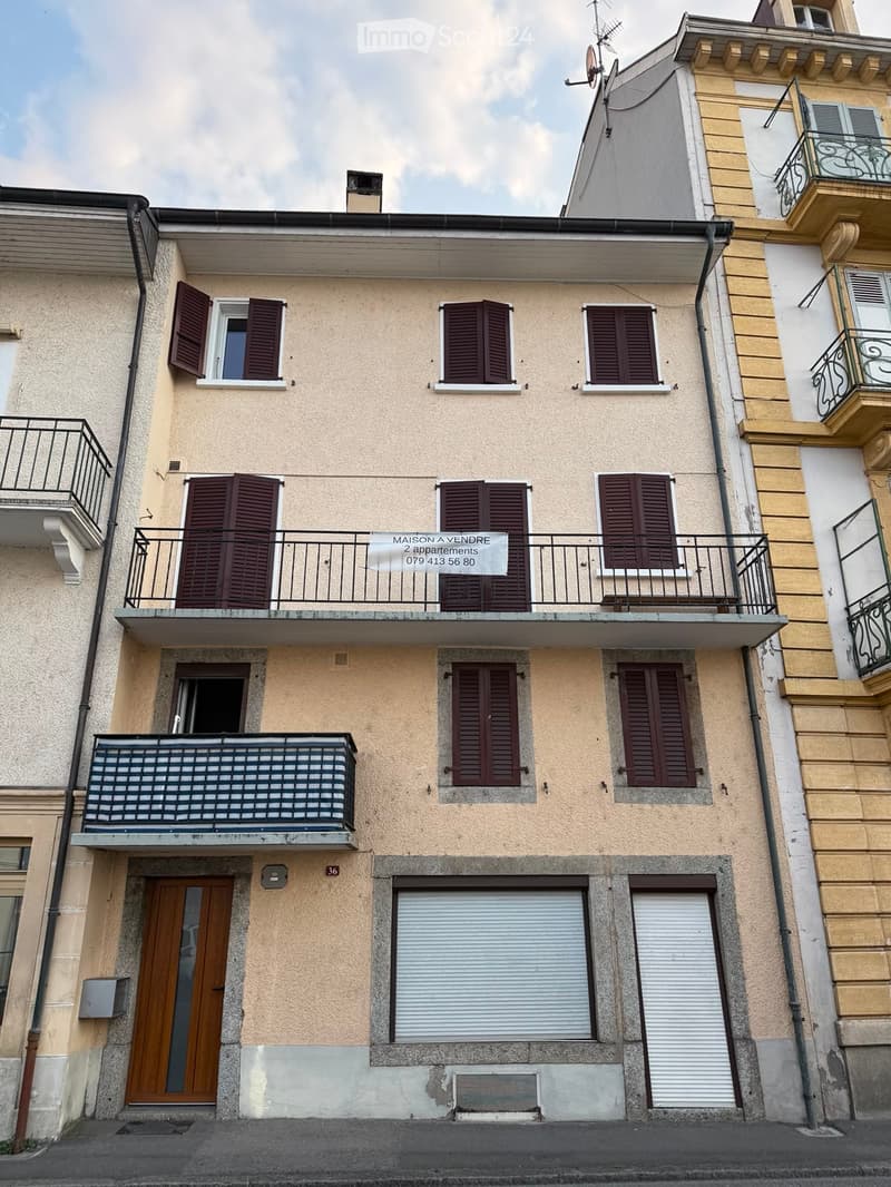Two-story residential building with beige facade, multiple windows with brown shutters, two balconies, and a garage entrance.