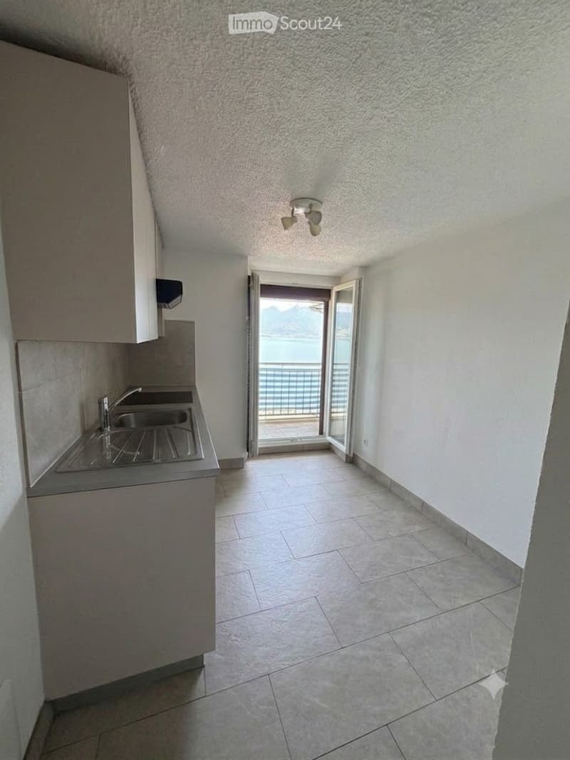 Modern kitchen with tiled floor, white walls, and a view of the sea through an open balcony door.
