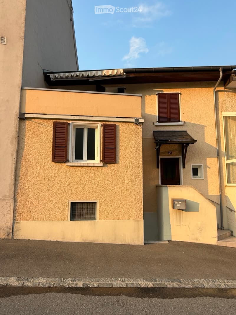 Two-story house with beige exterior, multiple windows with brown shutters, small balcony, and a covered entrance.