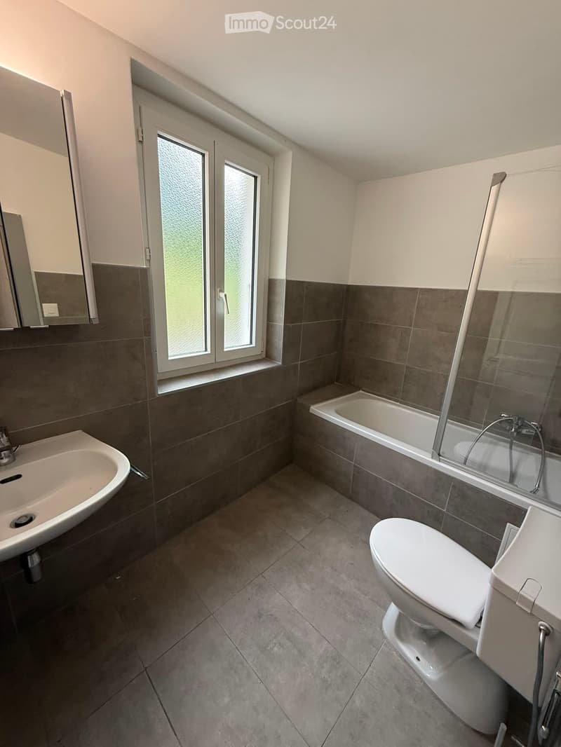 Modern bathroom with dark tiled walls, a white sink, toilet, bathtub, and a large window with frosted glass.