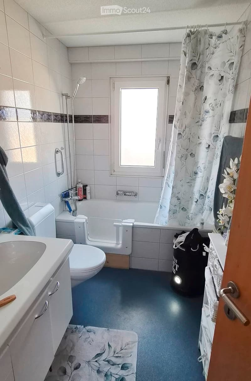 Bathroom with white tiles, a bathtub, toilet, sink, shower curtain, and a window. Blue floor and a floral rug are visible.