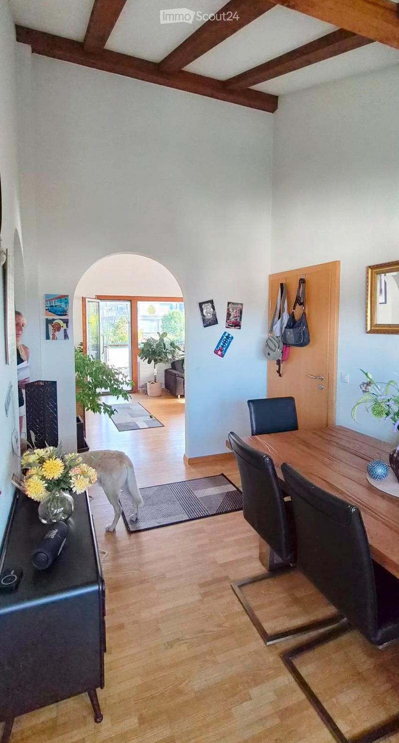Bright hallway with wooden flooring, a dining area with a wooden table and black chairs, a vase with yellow flowers on a sideboard, and a dog walking through the space.