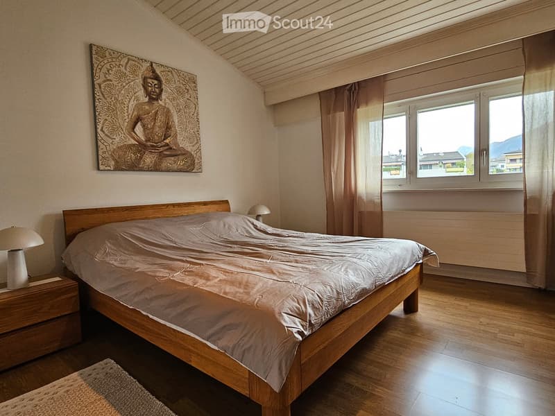 Cozy bedroom with wooden bed, nightstand, lamp, and a large painting on the wall. Large windows offer a scenic view.