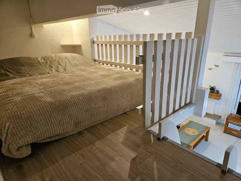 Cozy bedroom with a wooden bed, beige bedding, a small wooden stool, a small table with a screen, and a small plant on a shelf.