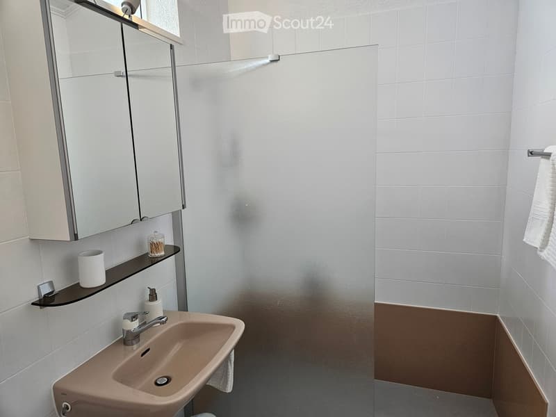Modern bathroom with a beige sink, chrome faucet, mirrored cabinet, and a black shelf holding a cup and toiletries. The walls are tiled in white with a brown lower section.