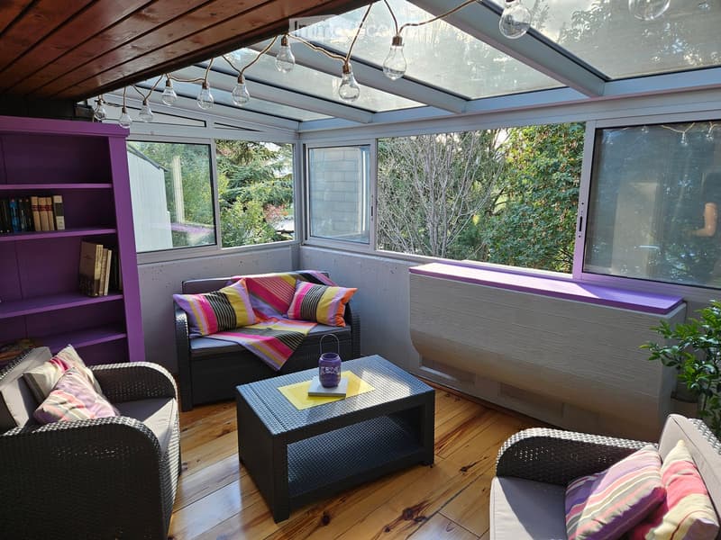 Brightly lit sunroom with wooden floors, large windows, and skylights. Features a wicker sofa with colorful pillows, a coffee table with a decorative item, a bookshelf, and additional seating.
