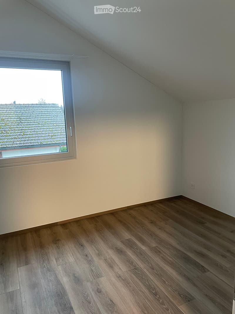 Empty room with wooden flooring and a large window offering a view of a roof. The walls are painted in a light color, and the room has a simple, clean design with no furniture or decorations.