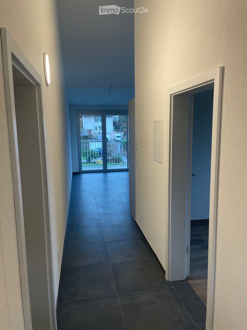 A clean, modern hallway with tiled flooring, white walls, and recessed lighting. Features include a sliding glass door leading to an outdoor area and several closed doors along the sides.
