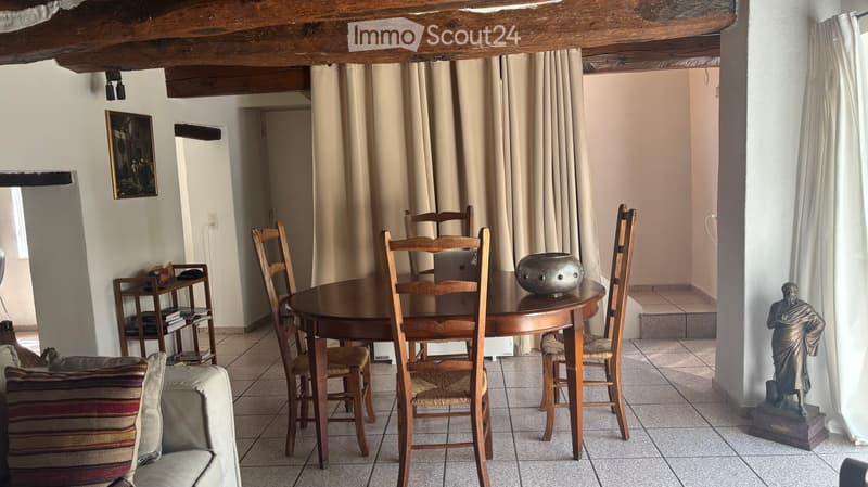 Cozy dining area with a round wooden table, six wooden chairs, a sofa with striped cushions, a small bookshelf, and a decorative statue on a pedestal.