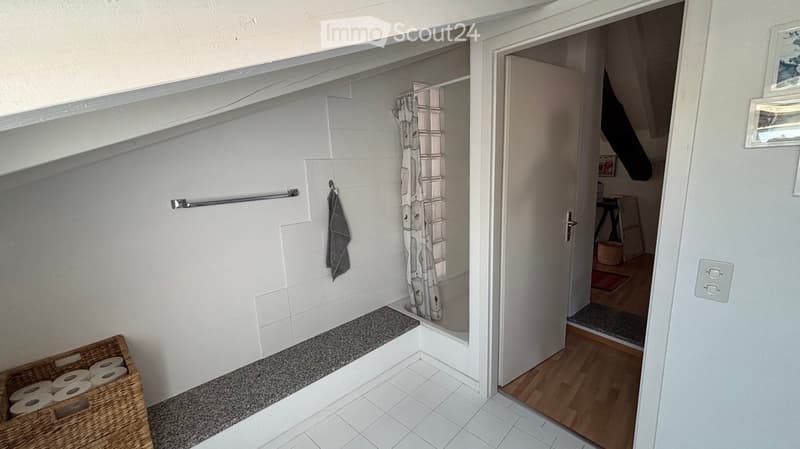 Modern bathroom with white tiles, a shower area with a curtain, a towel rack, and a basket of toilet paper. The adjacent room has wooden flooring and a small table.