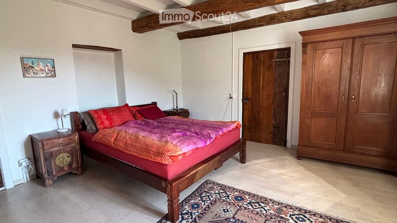 Cozy bedroom with wooden bed, colorful bedding, nightstand with lamp, large wooden wardrobe, and a patterned rug on the floor.