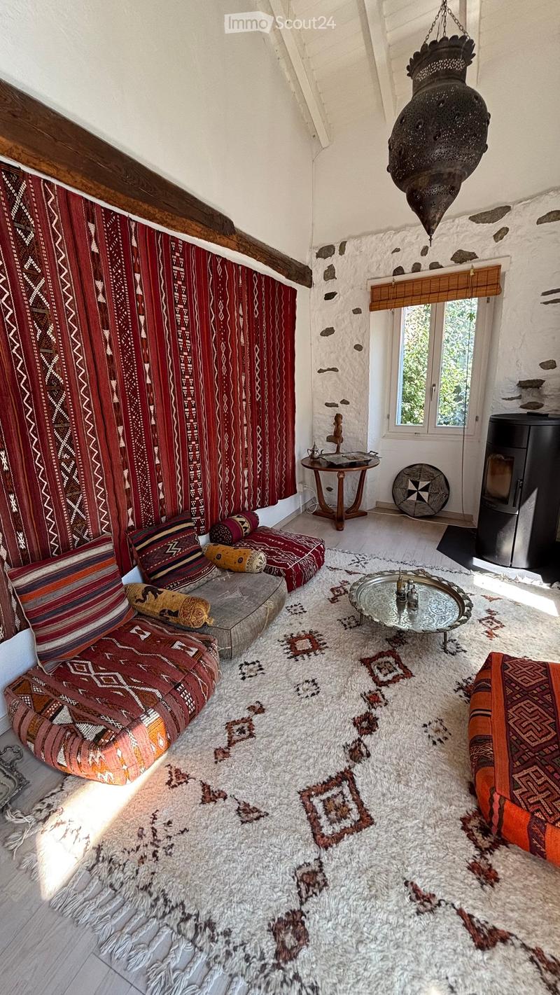 A cozy living room with a large, patterned rug, a low seating area with colorful cushions, a small wooden table, and a hanging decorative lantern. The room features exposed wooden beams and a stone wall with a window letting in natural light.
