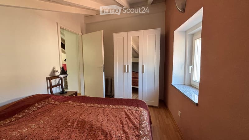Bedroom with a wooden floor, a bed with a patterned red bedspread, a white wardrobe, a small wooden chair with a table lamp, and a window with a small balcony.