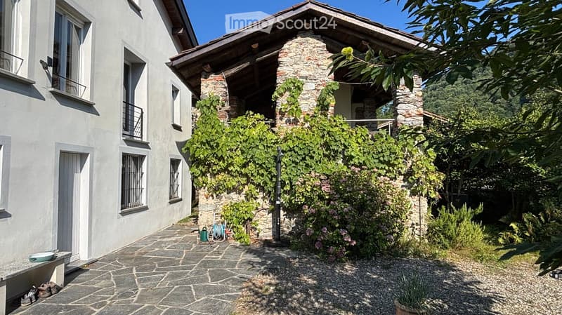 Two-story house with white exterior, stone chimney, and lush garden area with climbing plants and greenery.