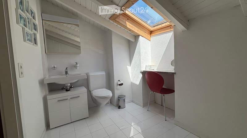Modern bathroom with white tiles, a skylight window, a sink with a cabinet, a toilet, a red chair, and a mirror above the sink.