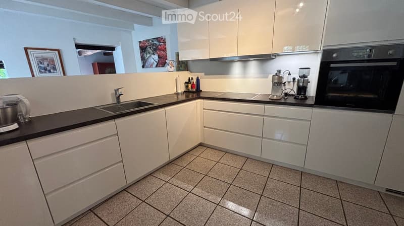 Modern kitchen with white cabinets, tiled floor, and various appliances including a sink, oven, coffee maker, and blender. There are also decorative items like a framed picture and a cup on the counter.