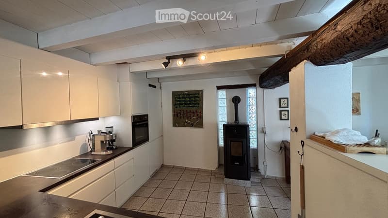 Modern kitchen with white cabinets, induction cooktop, oven, coffee maker, and a small fireplace. Exposed wooden beam on the ceiling and tiled floor.