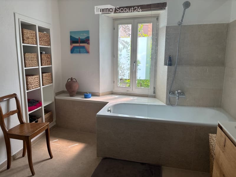 Bathroom with a large bathtub, wooden chair, shelf with woven baskets, vase, painting, and a shower area with a glass enclosure.
