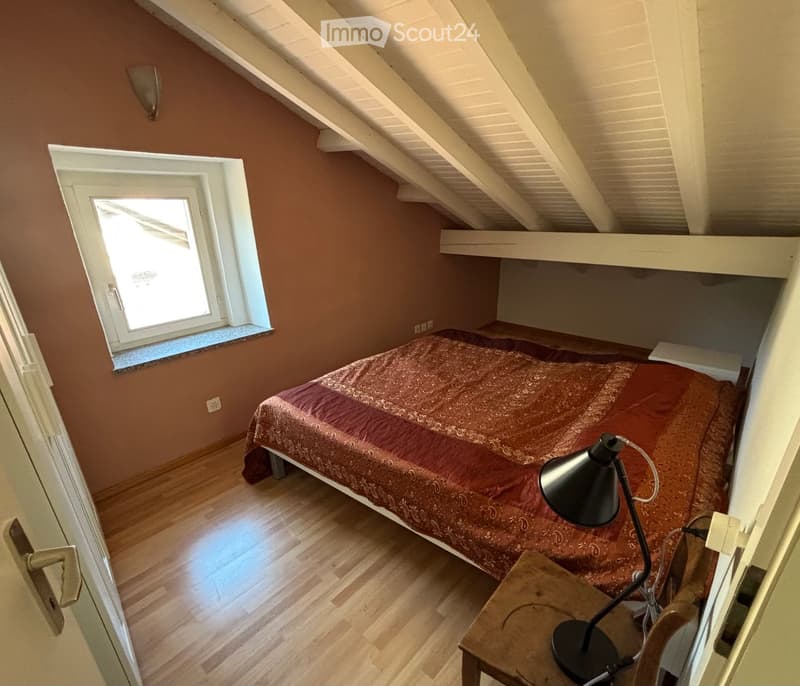 Cozy attic bedroom with wooden floors, slanted ceilings, a bed with patterned bedding, a table lamp on a nightstand, and a window letting in natural light.