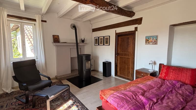 Cozy bedroom with exposed wooden beams, a bed with colorful bedding, a black armchair, a nightstand with a lamp, a modern fireplace, and several picture frames on the walls.