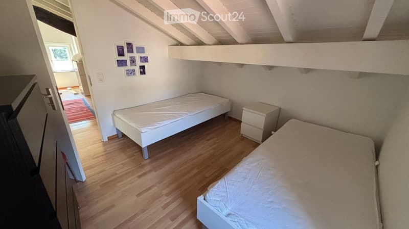 Cozy bedroom with two single beds, a nightstand, and a slanted wooden ceiling. The room features light wooden flooring and a small window with a view of greenery.
