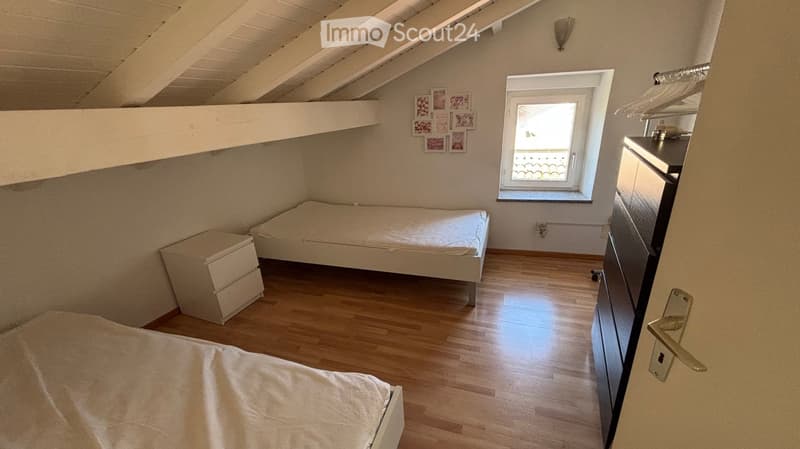 Cozy attic bedroom with wooden floors, slanted ceilings, a single bed, a nightstand, and a wardrobe. A window provides natural light.