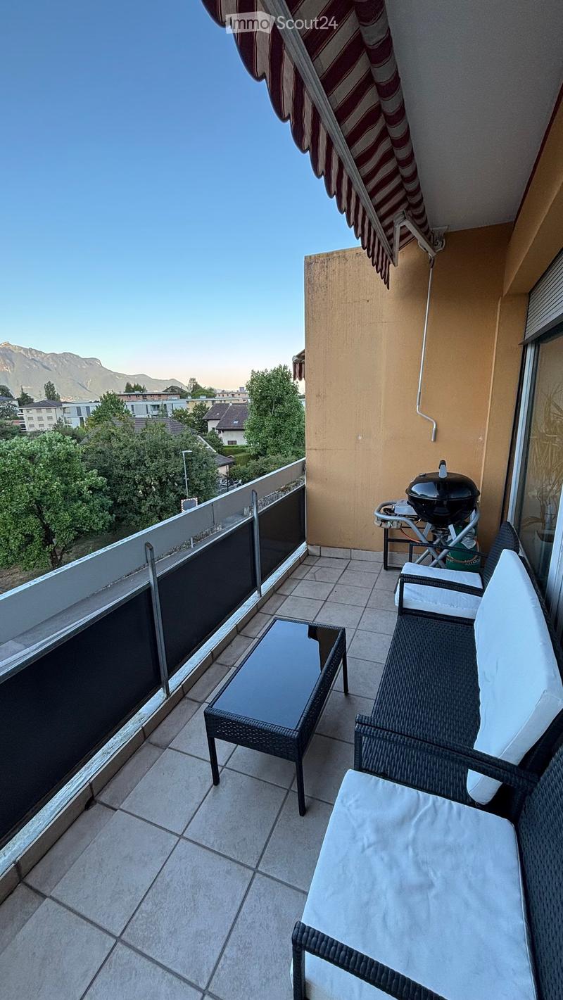 Balcony with tiled floor, glass railing, outdoor furniture including a table and chairs, and a view of the surrounding area with trees and mountains.