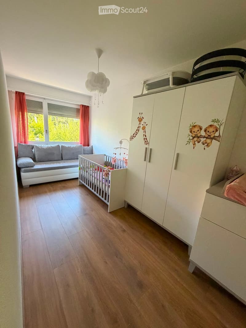 Brightly lit children's bedroom with wooden flooring, a white sofa, a baby cradle, a large wardrobe with animal decals, and a window with red curtains.