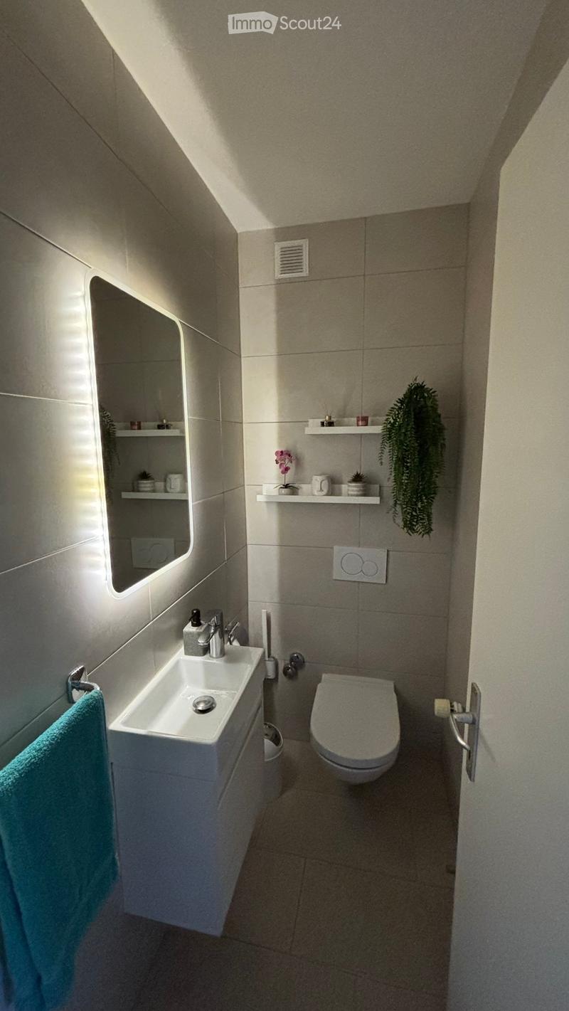 Modern bathroom with a white sink, toilet, mirror, shelves, and a small plant. The walls are tiled in a light gray color, and the floor is also tiled.