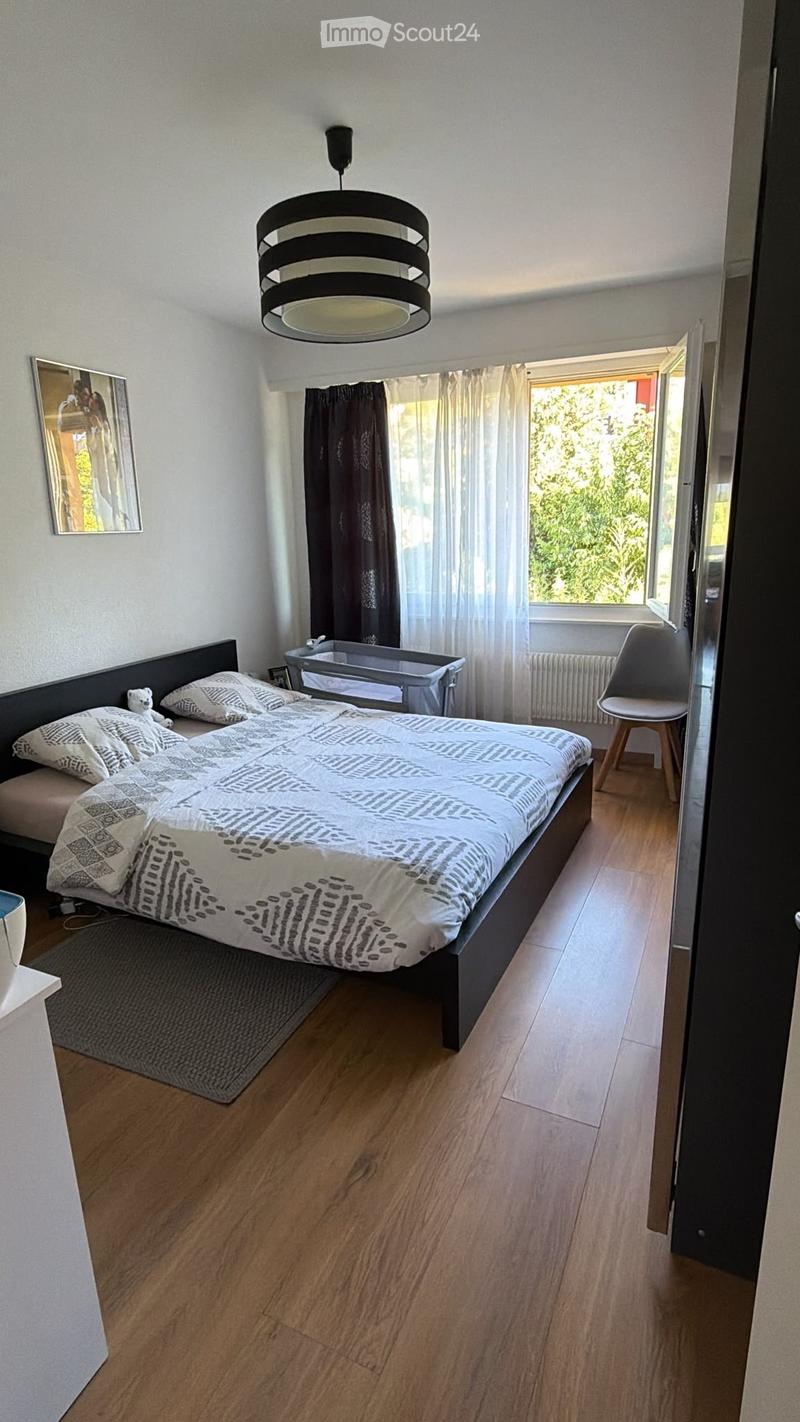 Modern bedroom with wooden flooring, large bed with patterned bedding, a baby crib, a chair, and a hanging lamp.