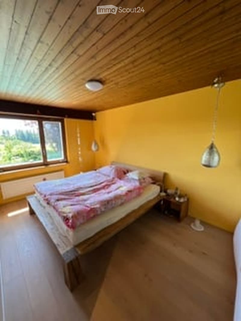 Bright bedroom with yellow walls, wooden ceiling, large windows offering a scenic view, a wooden bed with colorful bedding, a nightstand, and pendant lights.