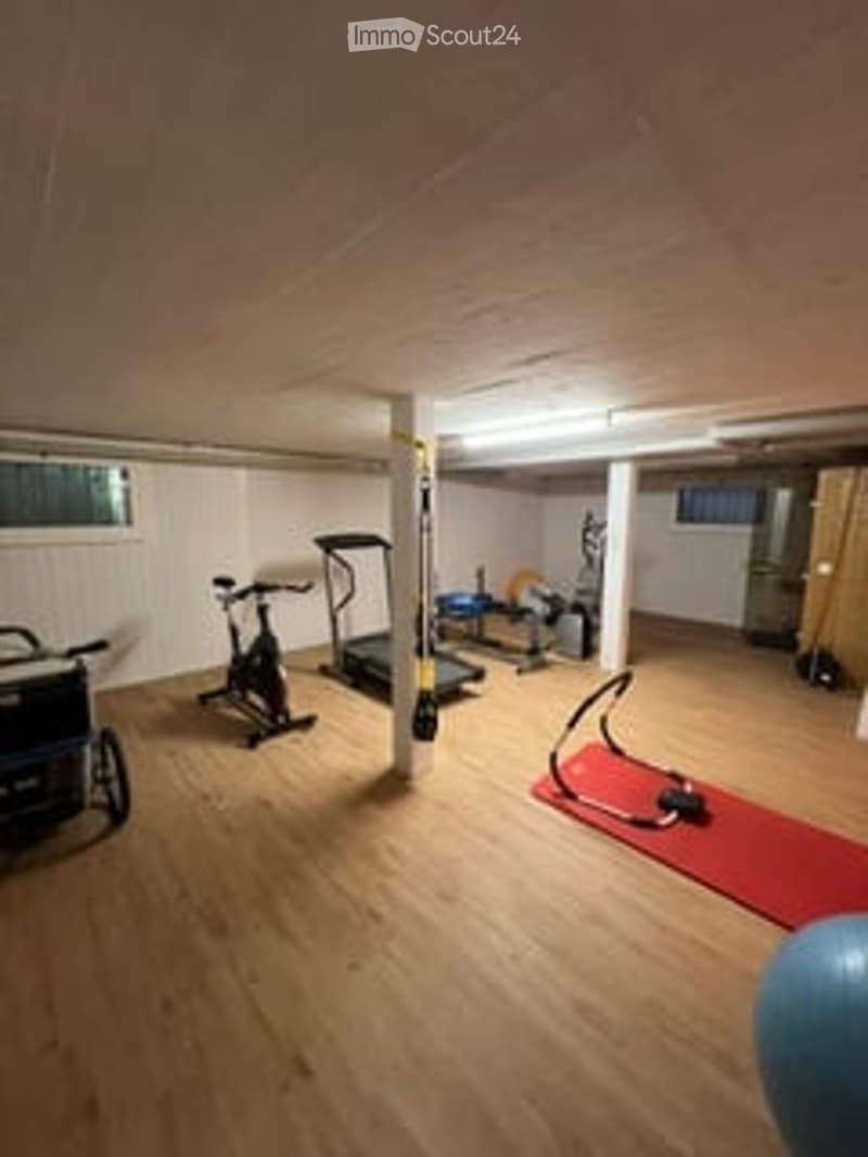 Well-lit basement gym with wooden flooring, exercise bike, treadmill, elliptical machine, weight machine, yoga mat, and exercise ball.