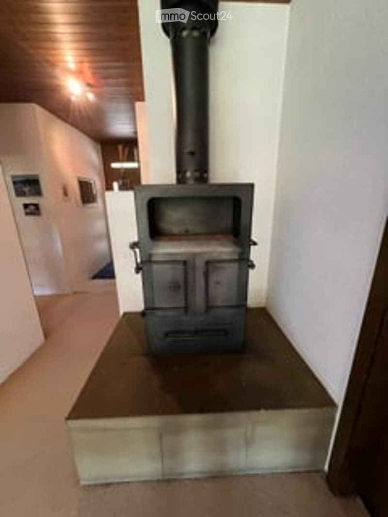 A wood-burning fireplace with a black chimney pipe, situated on a concrete platform in a room with white walls and a wooden ceiling.