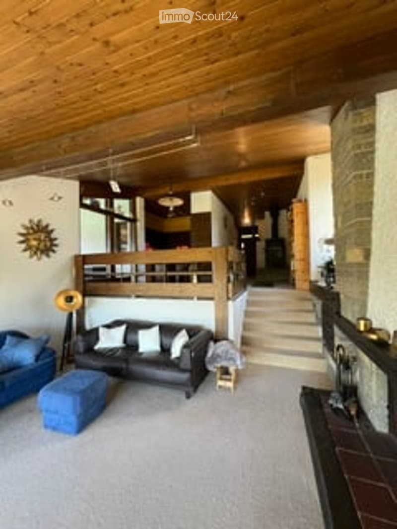 Spacious living room with wooden ceiling beams, fireplace, large windows, and comfortable seating including a sofa, ottoman, and armchair.