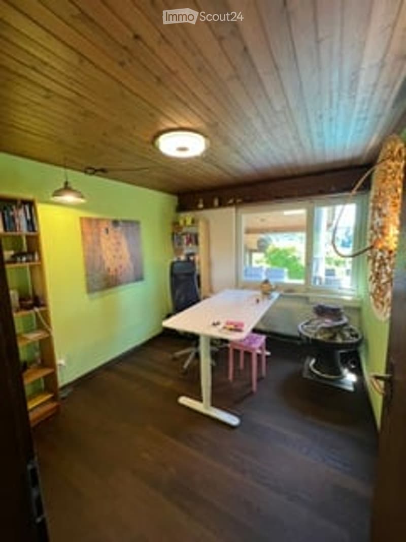 Bright room with wooden floors and ceiling, green walls, large window, table with chair, bookshelf, and radiator.
