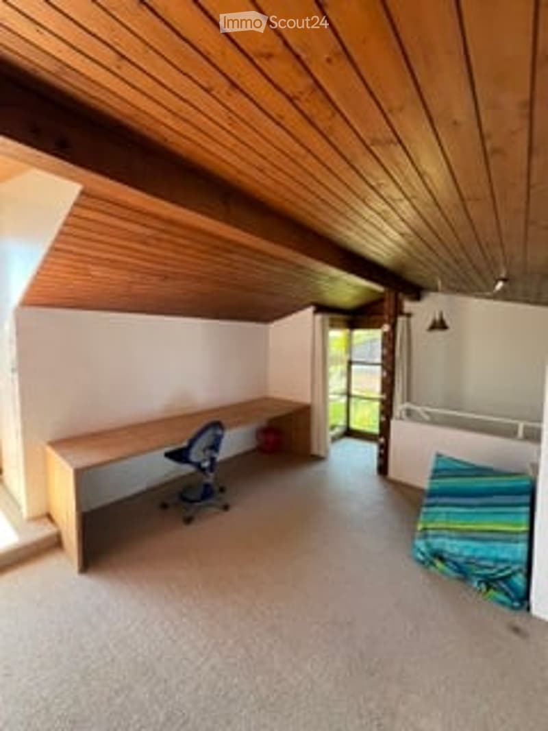 Bright room with wooden ceiling beams, a desk with a chair, and a bed with a colorful blanket.